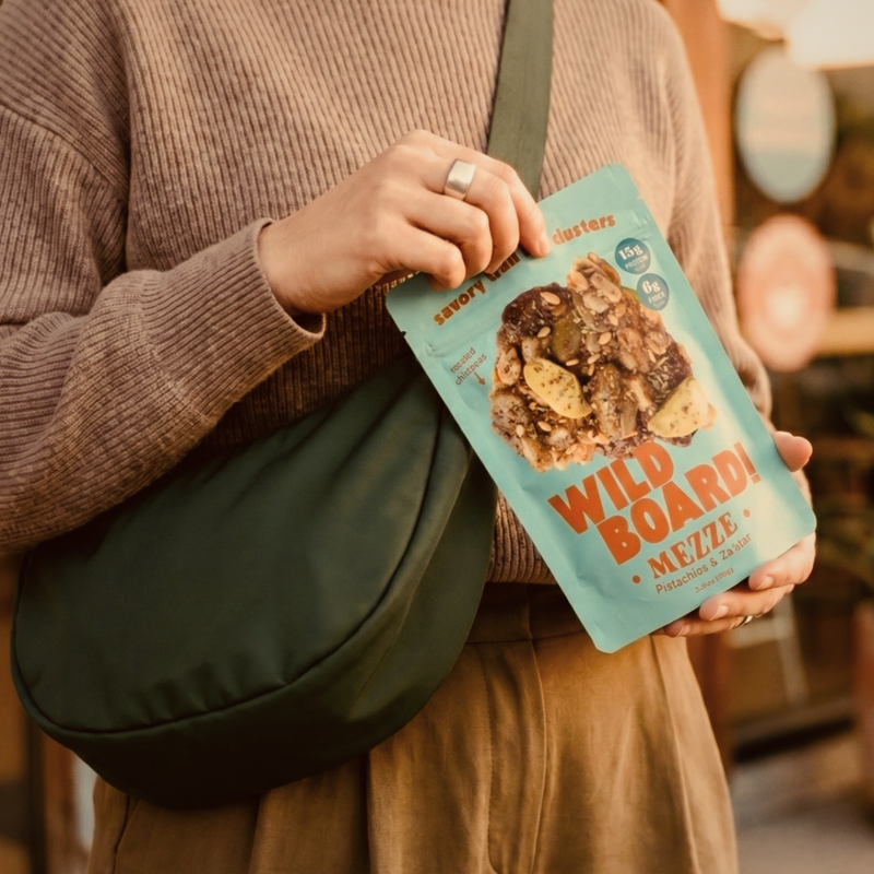 A woman with a crossbody bag holding a WildBoard Mezze pouch, savory trail mix clusters that need no refrigeration