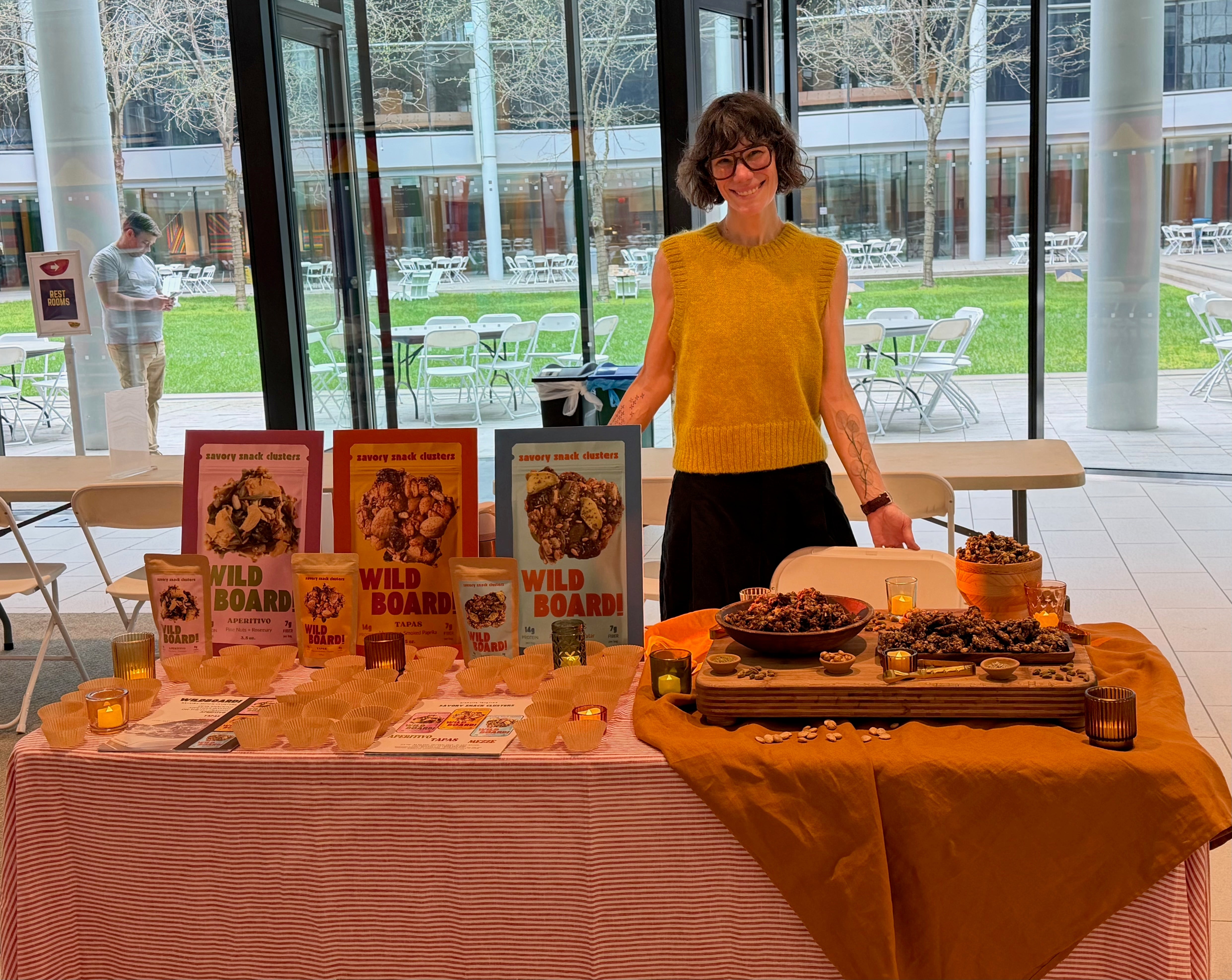 WildBoard founder Amy Boysen at a sampling table with savory trail mix cluster pouches, sample cups, and a tasting board of clusters
